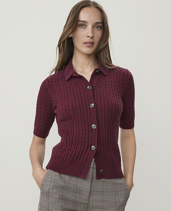A woman in a burgundy cable-knit polo shirt with her arms crossed stands against a plain white background.