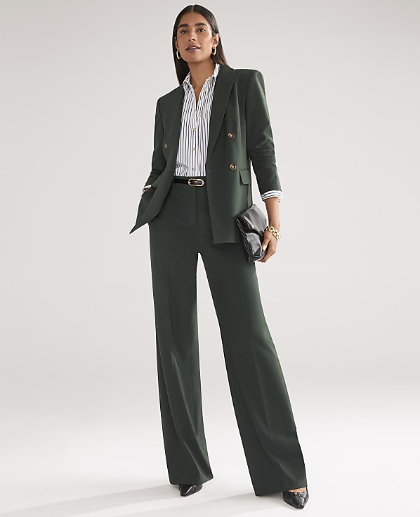 A woman in a dark green pantsuit stands against a plain, light gray background, holding a black clutch.