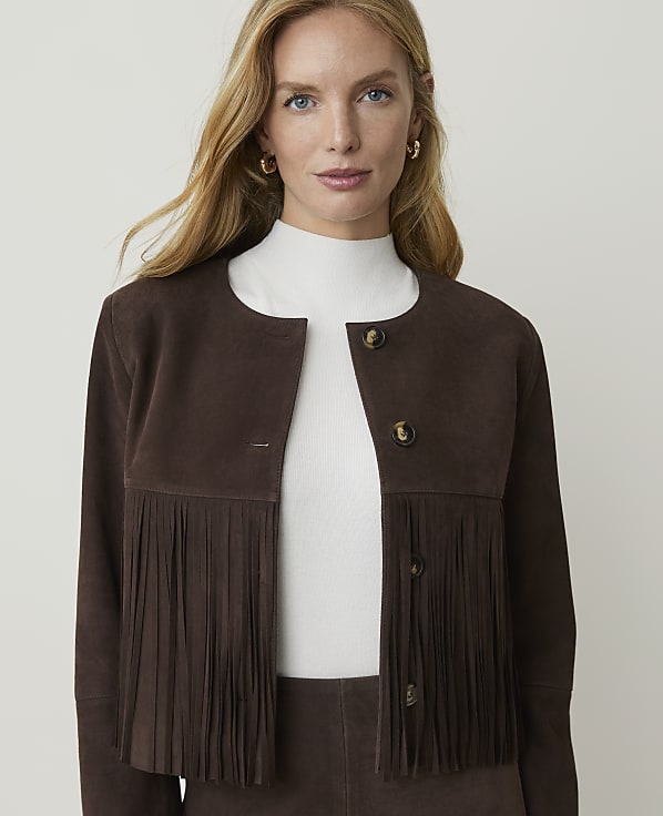 A woman with blonde hair and blue eyes, wearing a brown suede jacket with fringe, stands against a plain white background.