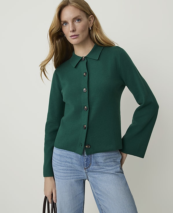 A woman in a green collared cardigan and blue jeans stands against a plain white background.