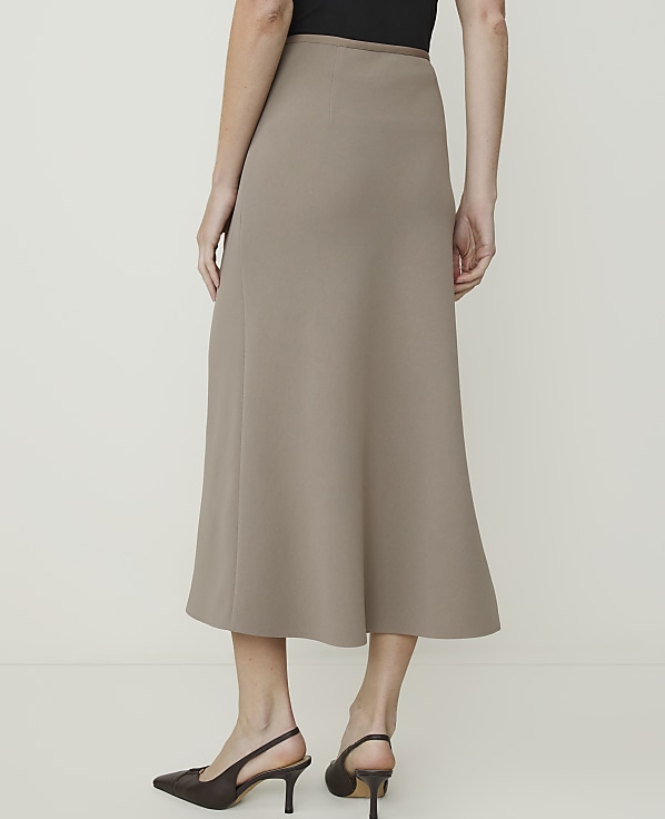 A woman in a taupe midi skirt and dark heels stands against a plain, light-colored background.