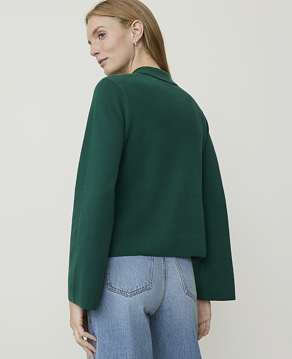 A woman in a green sweater and blue jeans stands against a plain, light background.