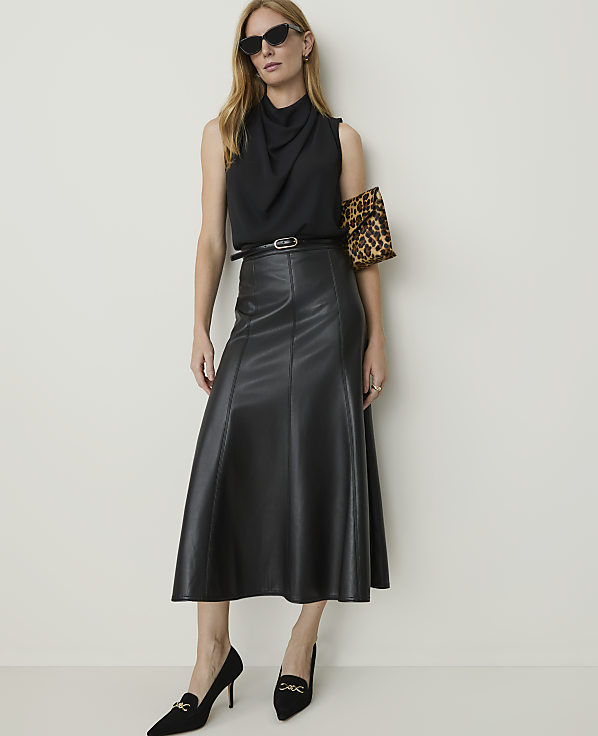 A woman in a black cowl-neck top and a black leather midi skirt poses against a plain white background, accessorized with sunglasses, a leopard-print clutch, and black heels.