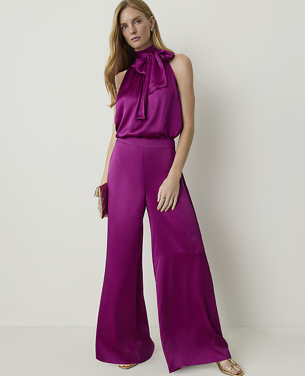 A woman in a magenta halter-neck jumpsuit with wide-leg pants stands against a plain, light-colored background, holding a matching clutch.