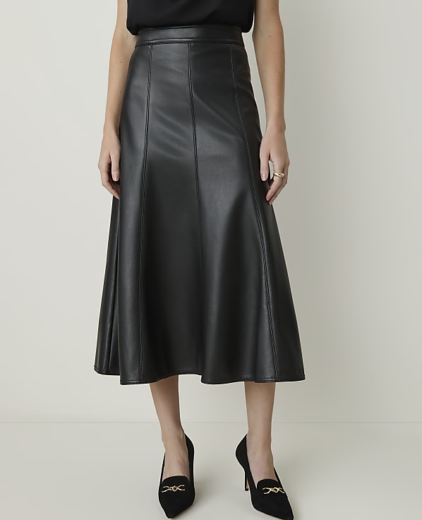 A woman in a black faux leather midi skirt and black heels stands against a plain, light-colored background.