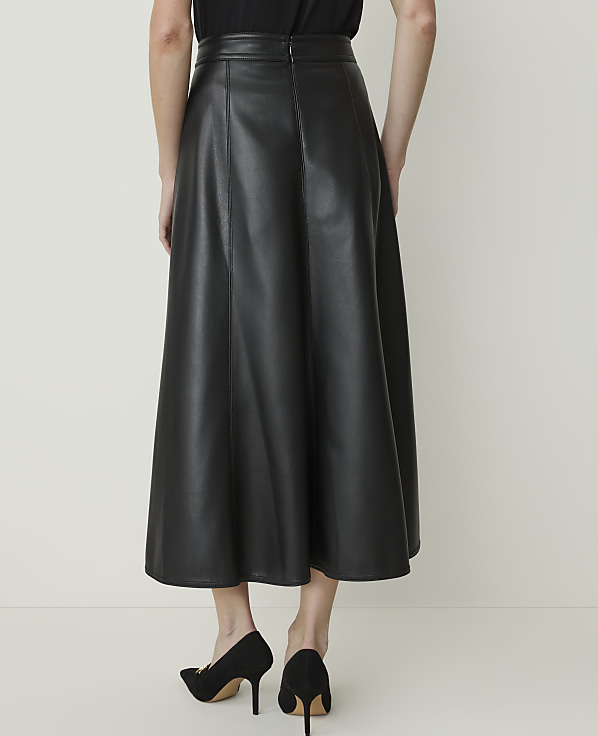 A person wearing a black leather midi skirt and black heels stands against a plain, light-colored background.