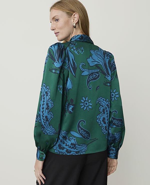A woman in a green paisley blouse stands against a plain, light gray background.