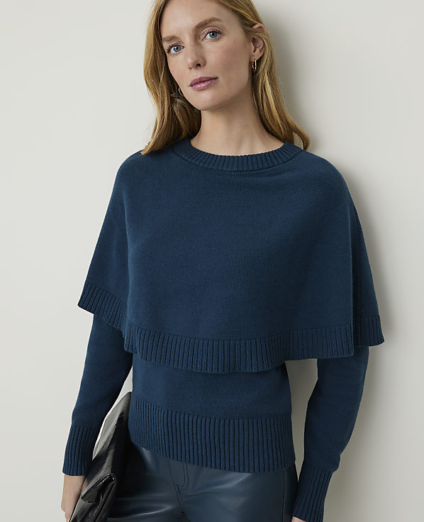 A woman in a teal sweater with a capelet and dark pants holds a black clutch against a plain background.