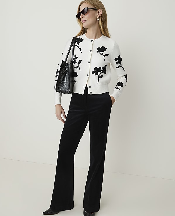 A woman in a floral cardigan and black flared pants stands against a plain white background.