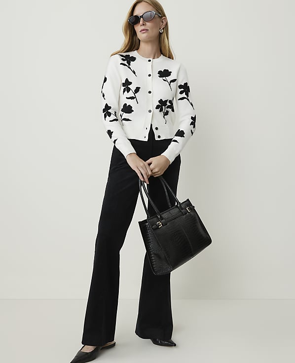 A woman in a floral cardigan and black flared pants poses with a black handbag against a plain white background.