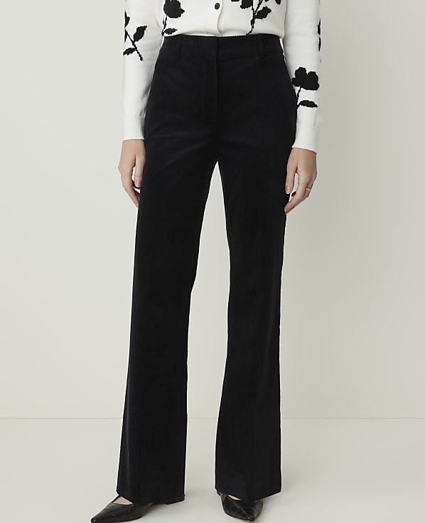 A person stands in black corduroy flared pants and a white floral sweater against a plain white background.