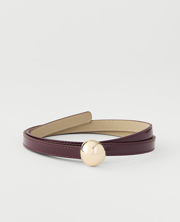 A thin, glossy burgundy belt with a gold spherical buckle is coiled on a plain white background.