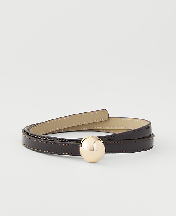 A thin, dark brown leather belt with a gold sphere buckle is coiled on a plain white background.