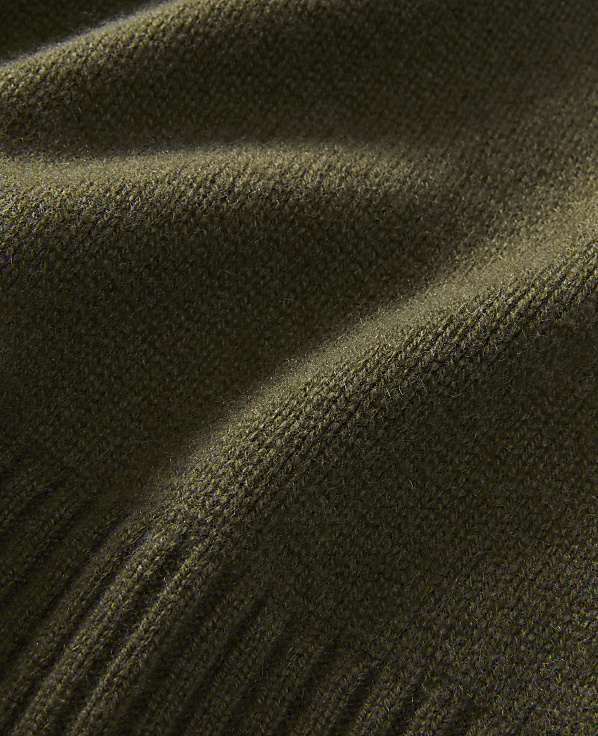 A close-up of a textured olive green knitted fabric fills the frame, with soft folds and a ribbed edge visible in the foreground and a smooth, slightly blurred expanse in the background.