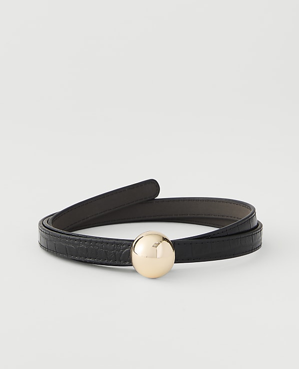 A black, textured belt with a gold sphere buckle is coiled on a plain white background.