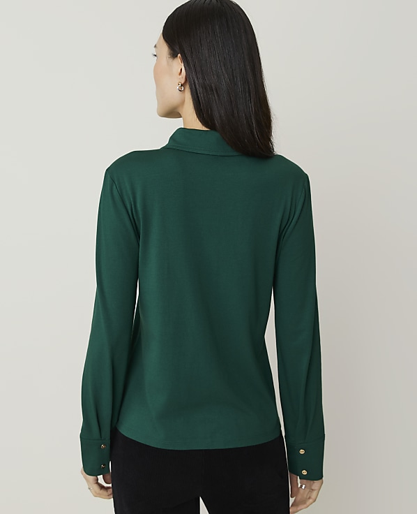 A woman in a green collared shirt stands against a plain, light gray background.
