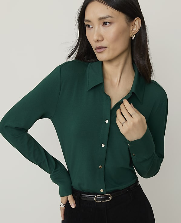 A woman in a green collared shirt stands against a plain, light gray background.
