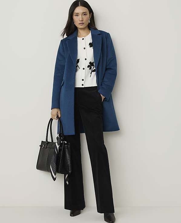 A woman in a teal coat and black pants stands against a plain white background, holding a black handbag.