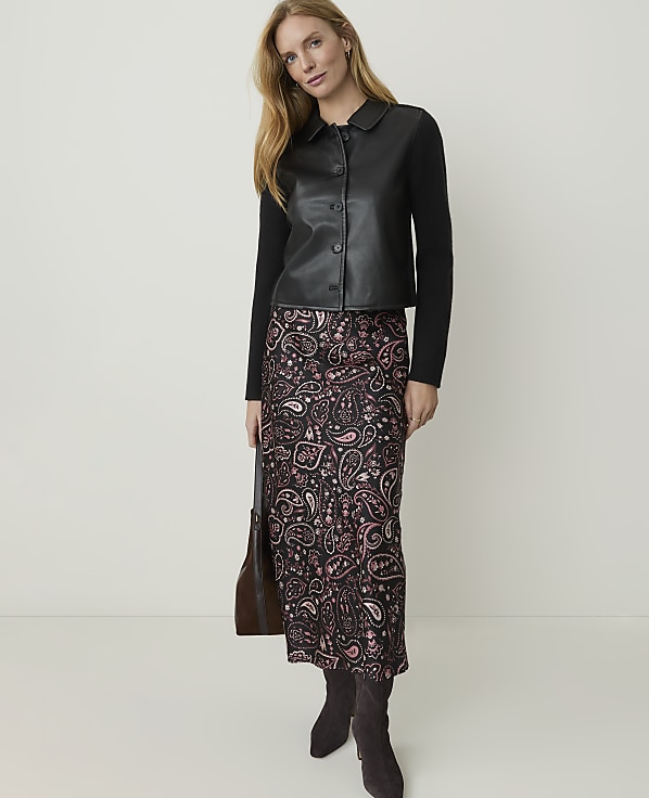 A woman in a black leather jacket and a patterned skirt stands against a plain white background, holding a brown handbag.
