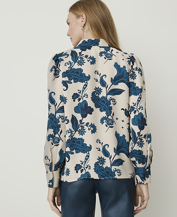 A woman in a cream-colored blouse with a blue floral pattern stands against a plain white background.