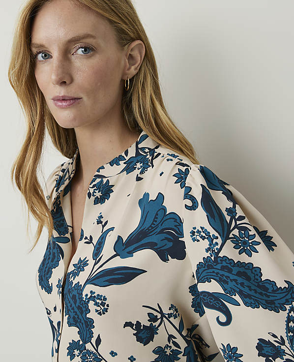A woman with blonde hair and fair skin wears a cream-colored blouse with a blue floral pattern, set against a plain, light background.