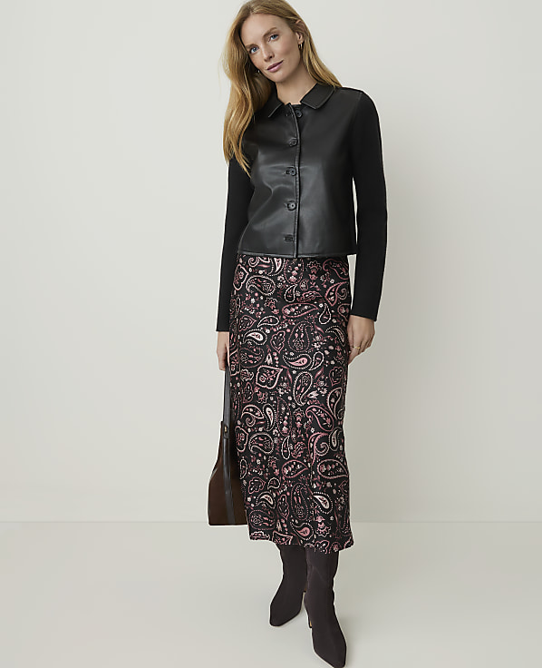 A woman in a black leather-look jacket and a patterned skirt stands against a plain, light-colored background.