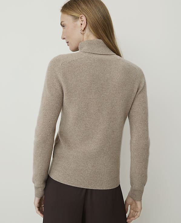 A woman in a beige turtleneck sweater stands against a plain, light-colored background.