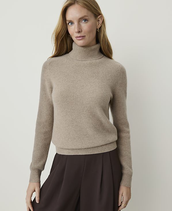 A woman in a beige turtleneck sweater and brown pants stands against a plain, light background.