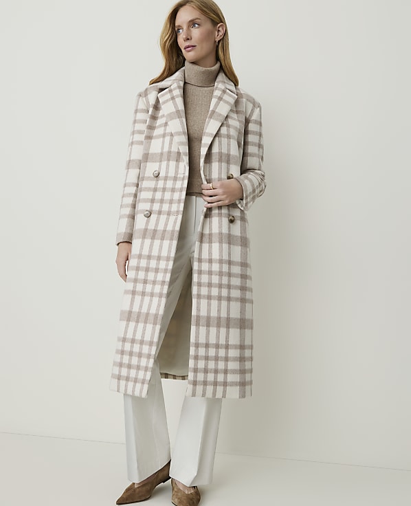A woman in a beige and white plaid coat and white pants stands against a plain, light background.
