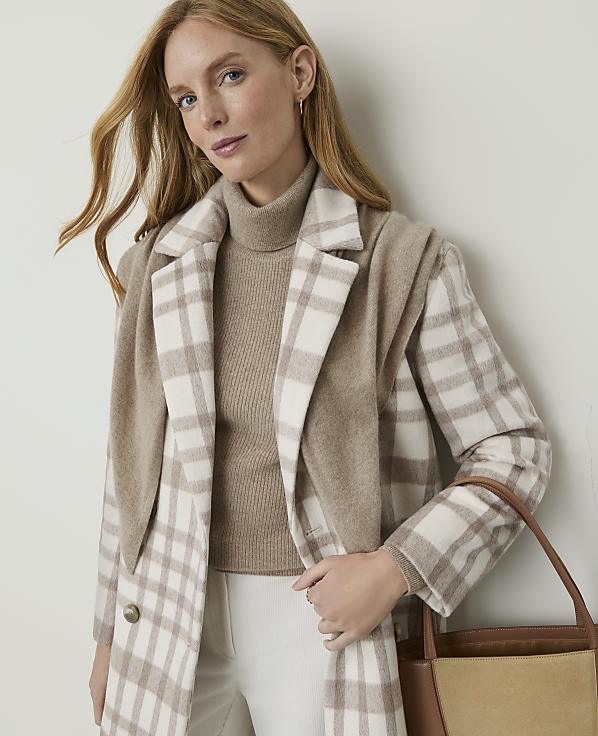 A woman in a beige turtleneck and a plaid coat stands against a plain, light-colored wall, holding a brown tote bag.