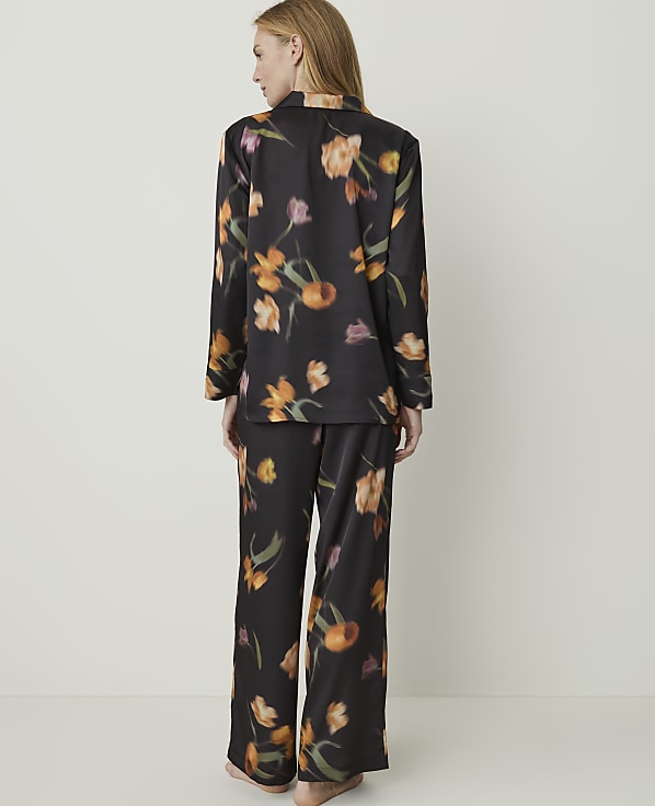 A woman stands facing away from the camera wearing black pajamas with a floral print, set against a plain white background.