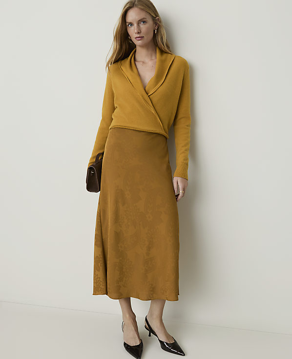 A woman in a mustard yellow wrap sweater and a patterned brown skirt stands against a plain cream background, holding a dark brown clutch.