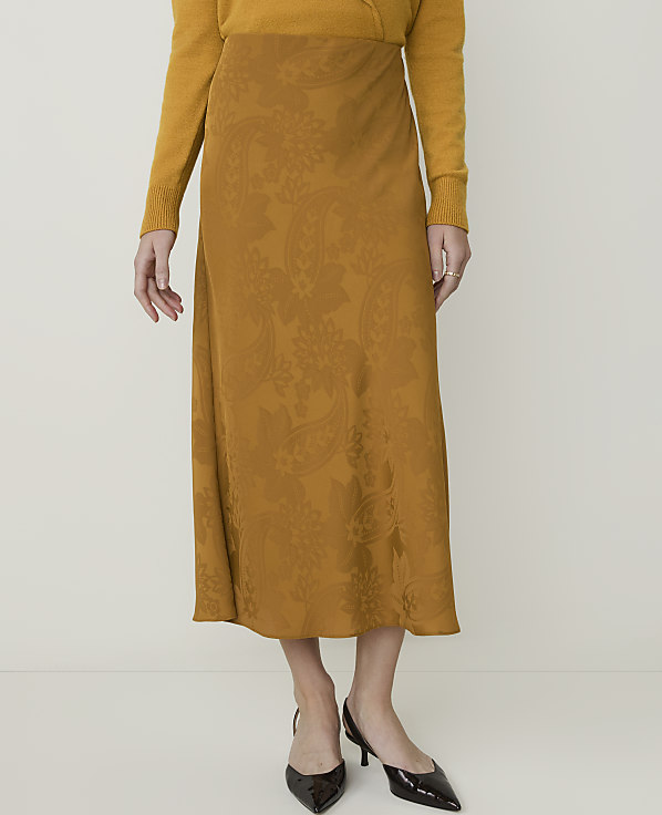 A woman in a mustard yellow sweater and a patterned midi skirt stands against a plain, light background.