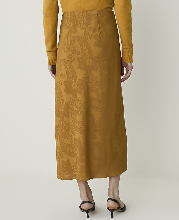 A person stands in a studio setting, wearing a patterned mustard-colored skirt and a matching sweater, with their back to the camera.