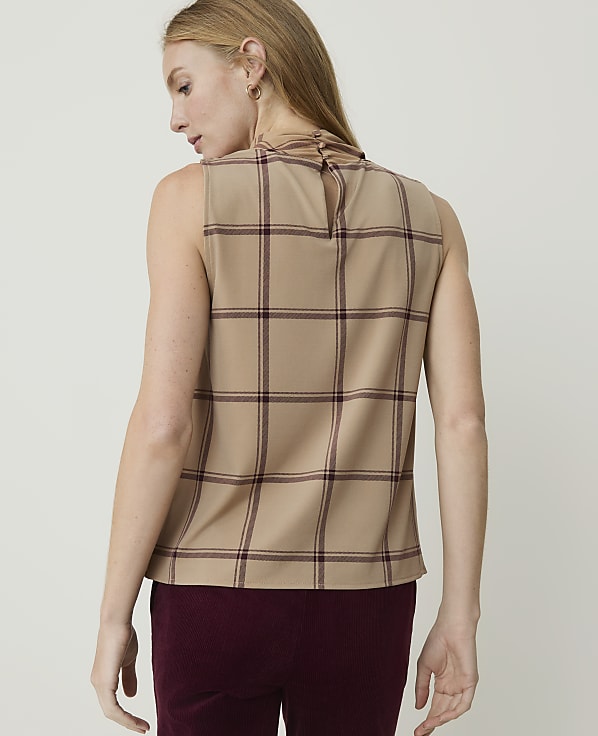 A woman in a tan plaid sleeveless top and burgundy pants stands against a plain white background.