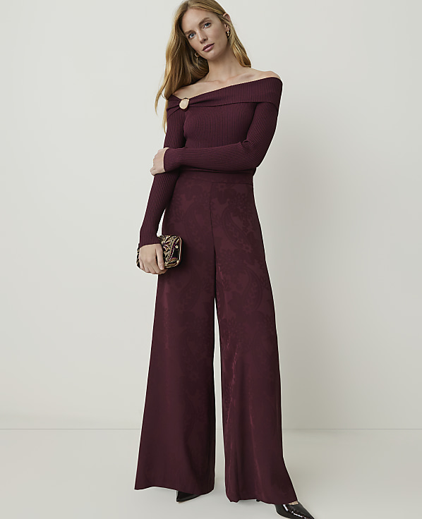 A woman in a burgundy off-the-shoulder jumpsuit and patterned clutch stands against a plain, light-colored background.