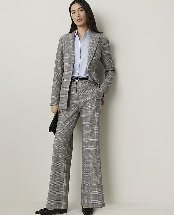 A woman in a plaid suit and blue shirt stands against a plain white background, holding a black clutch.
