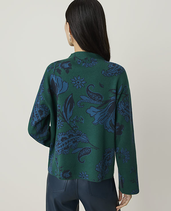 A woman in a green, patterned sweater and dark pants stands against a plain, light background.