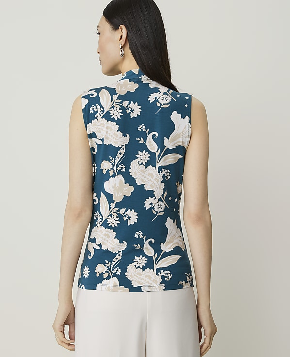 A woman in a teal floral sleeveless top stands against a plain white background.