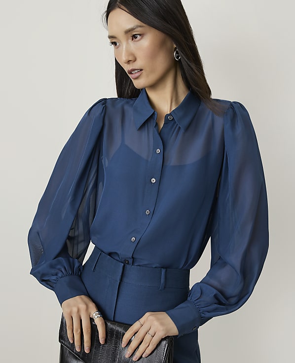 A woman in a sheer blue blouse and matching pants holds a black clutch against a plain background.