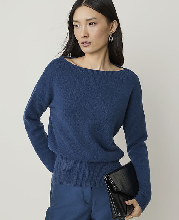 A woman in a blue sweater and pants holds a black clutch in front of a plain background.
