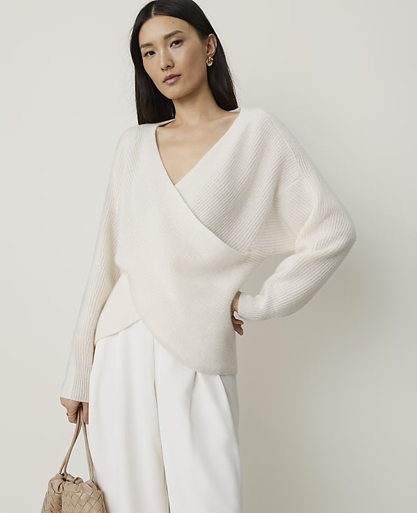 A woman in a cream wrap sweater and white pants stands against a plain, light background, holding a woven bag.