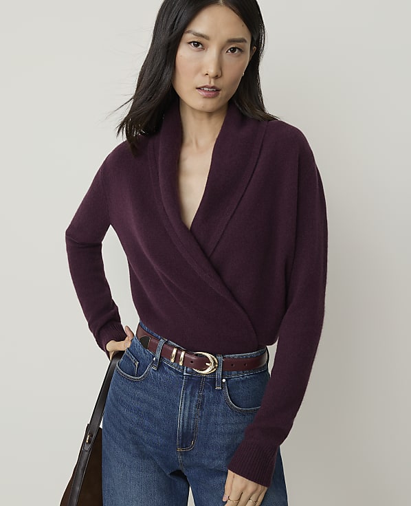 A woman in a plum-colored wrap sweater and blue jeans stands against a plain, light gray background.