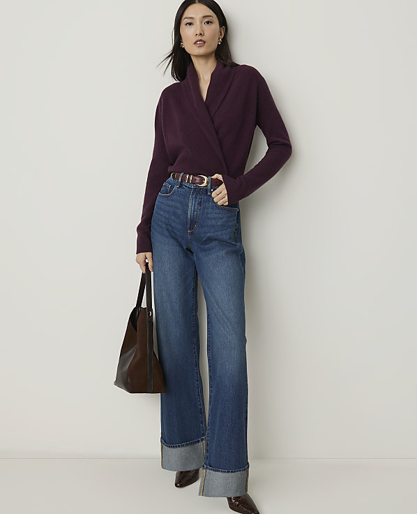 A woman in a plum-colored wrap sweater and wide-leg jeans stands against a plain white background, holding a brown leather tote bag.