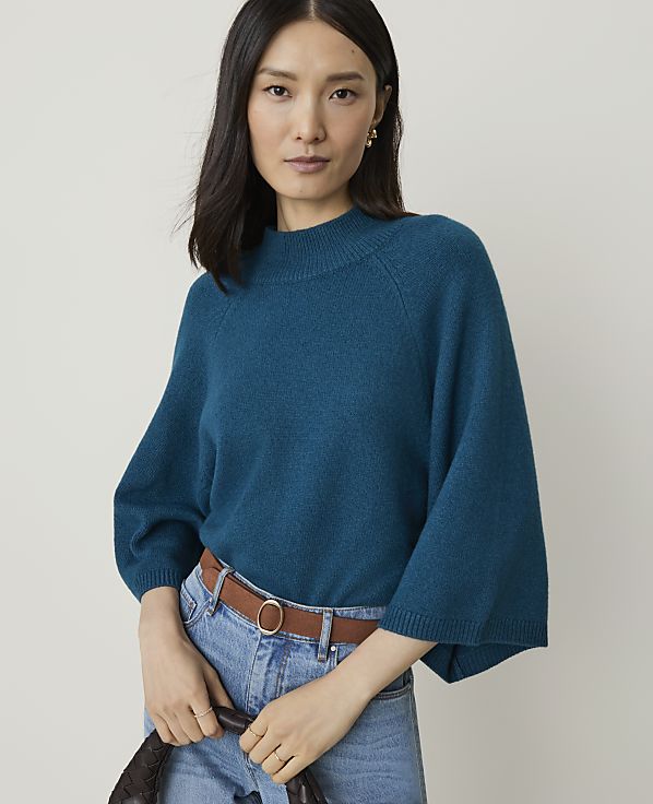 A woman in a teal sweater and jeans stands against a plain background.