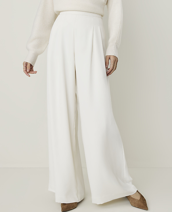 A person in a cream sweater and white wide-leg pants stands against a plain, light background.