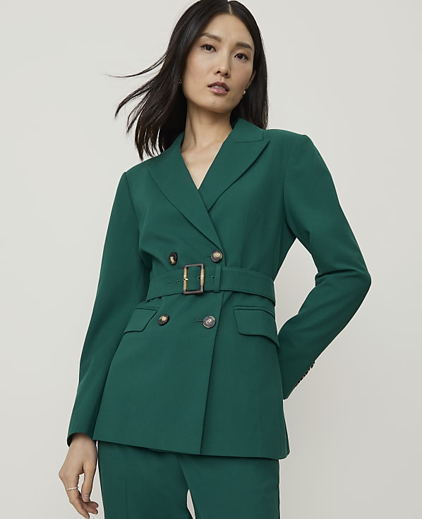 A woman in a green double-breasted blazer with a belt stands against a plain, light gray background.