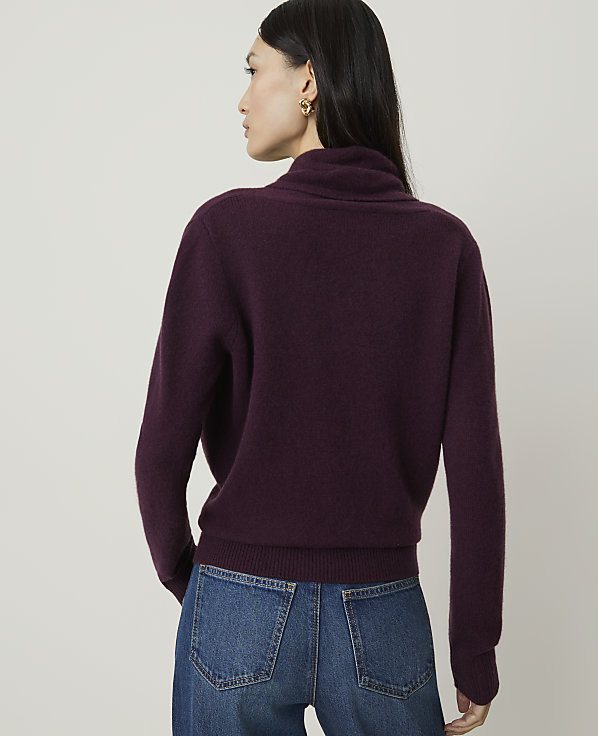 A woman in a burgundy turtleneck sweater and blue jeans stands against a plain, light gray background.