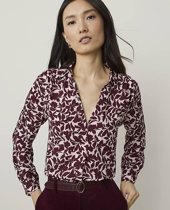 A woman in a floral blouse and maroon pants stands against a plain, light background.