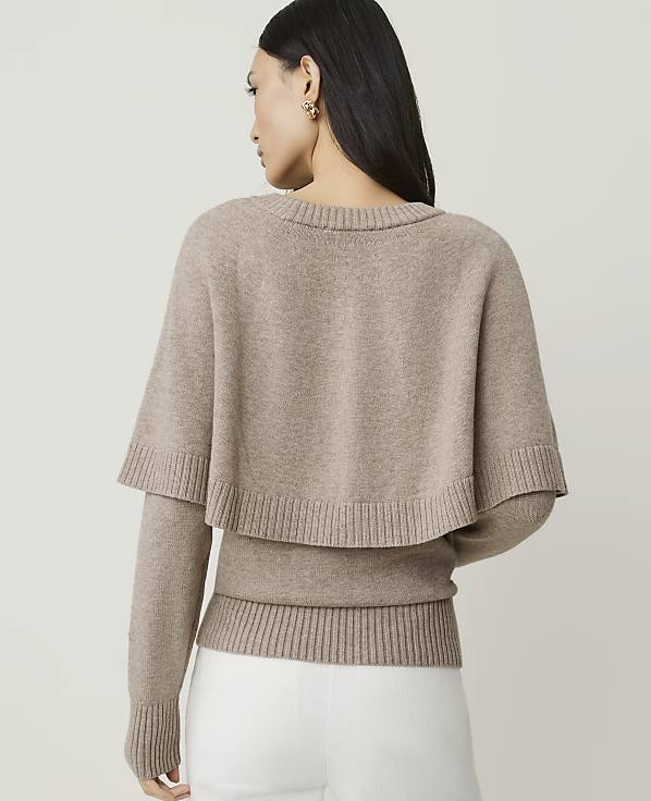 A woman in a beige sweater with a capelet stands against a plain, light background.